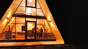 A triangular wooden cabin with a glass door entrance, a kitchen area with a dining table, and a visible fireplace. - Outscape Cerro Castillo (Río Ibáñez)
