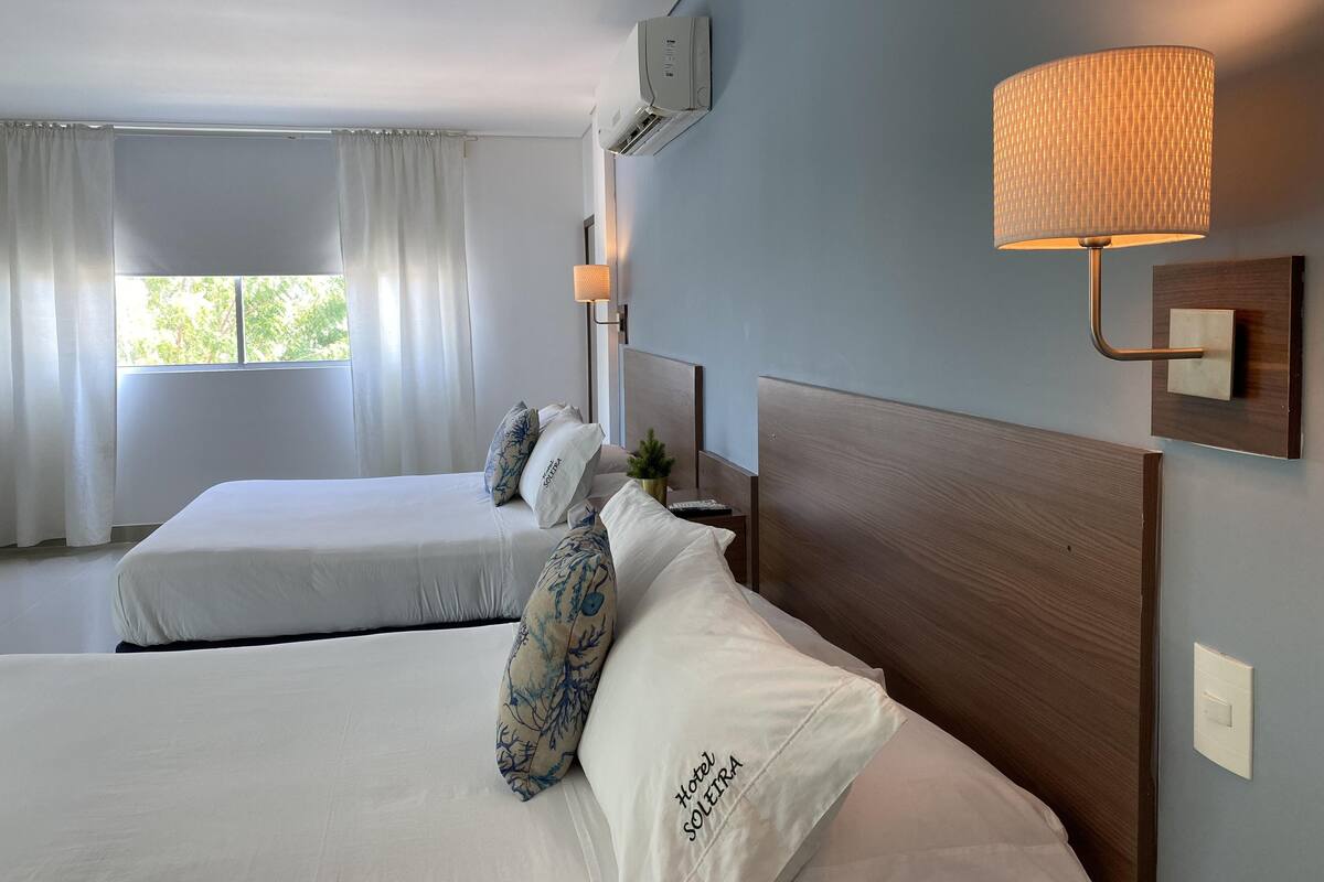 A hotel room with two beds, a wall-mounted lamp, and a window with curtains.