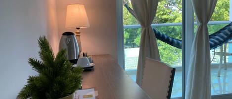 Hotelkamer met een houten tafel, een lamp, een potplant en uitzicht op het omliggende landschap door grote ramen met gordijnen.