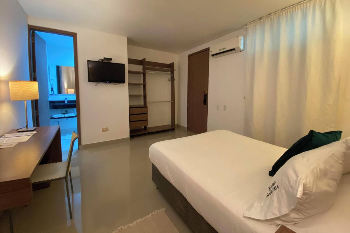 A hotel room with a bed, a desk with a lamp, a TV, a wardrobe, and a bathroom visible through an open door.