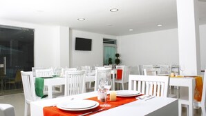 Un espace repas moderne avec des tables et des chaises blanches, un téléviseur à écran plat fixé au mur et une grande fenêtre laissant entrer la lumière naturelle.