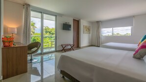 Une chambre d’hôtel avec un grand lit, un bureau avec une lampe, une télévision et un balcon donnant sur un espace verdoyant.