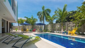 Une piscine entourée de palmiers et bordée d’un bâtiment moderne, avec des chaises longues et des parasols à proximité.