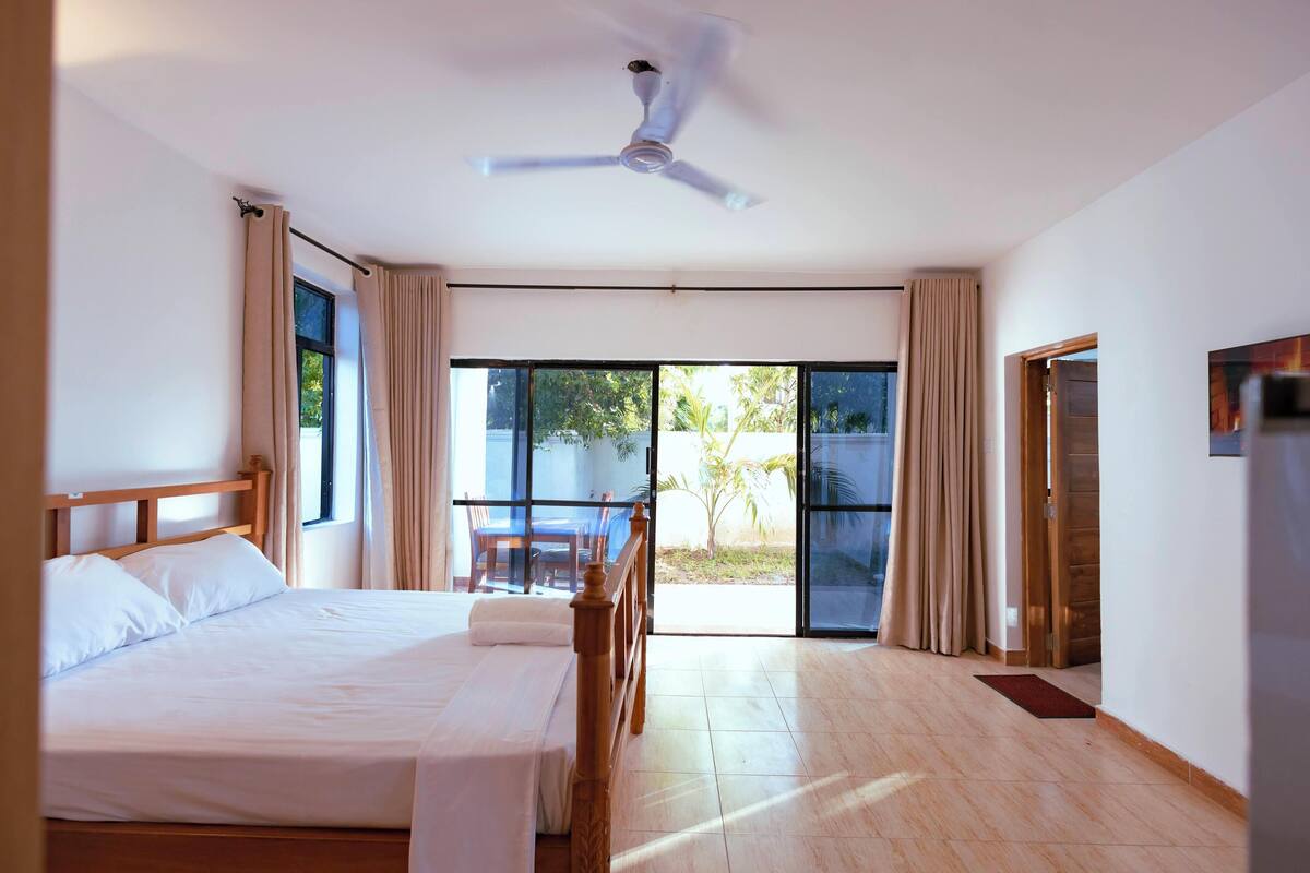 Ein Hotelzimmer mit einem Bett, einem Deckenventilator, Blick durch eine Schiebetür und einem Essbereich mit Tisch und Stühlen.