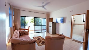 A living room with a sofa, armchairs, a flat-screen TV, a ceiling fan, and a sliding glass door leading to a balcony. - Diani Luxe Villa (Diani Beach)