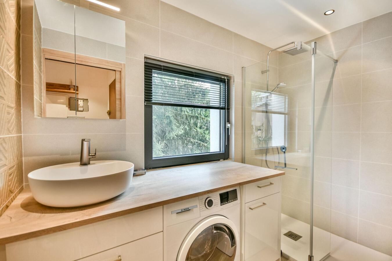 A modern bathroom with a large vessel sink, a Bosch washing machine, a glass shower enclosure, and a window with blinds.