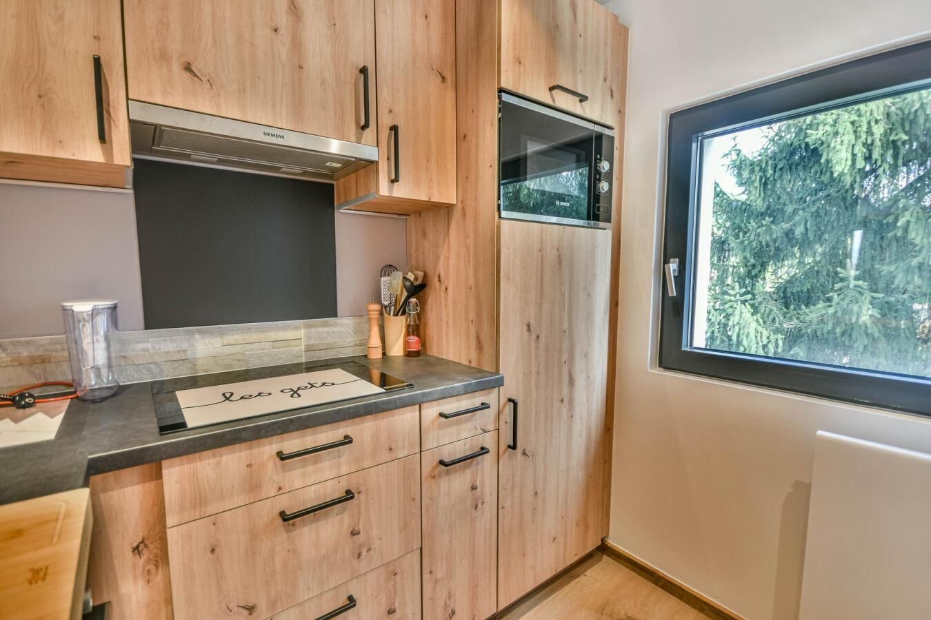 Une cuisine moderne avec des meubles en bois, une hotte de cuisinière en acier inoxydable, un four à micro-ondes Bosch et une fenêtre donnant sur les arbres.