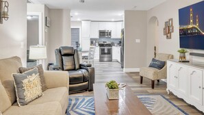 A modern living room with a leather sofa, a coffee table, and a TV. There is a kitchen in the background with white cabinets and a microwave.