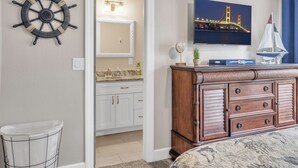 A bedroom with a bed, a dresser, a mirror, a wall-mounted ship's wheel, a television displaying a cityscape, and a small sailboat model.