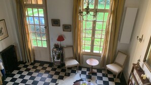 Ein Zimmer mit Schachbrettboden, zwei mit Vorhängen versehene Fenster, einer Kandelaber, ein kleiner Tisch mit einer Vase und eine Heizung.