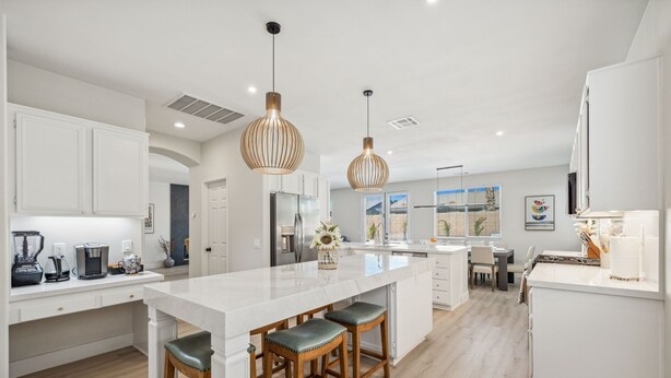 A modern kitchen with white cabinetry, a large island with bar stools, pendant lighting, and a dining area with a view. - NEW Lush Luxury Living-Heated Pool/Spa Included. Walking distance to Coachella (Indio)