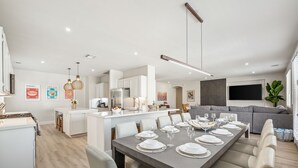 A modern dining area with a long table set for six, a kitchen in the background, and a grey sofa in the living area. - NEW Lush Luxury Living-Heated Pool/Spa Included. Walking distance to Coachella (Indio)