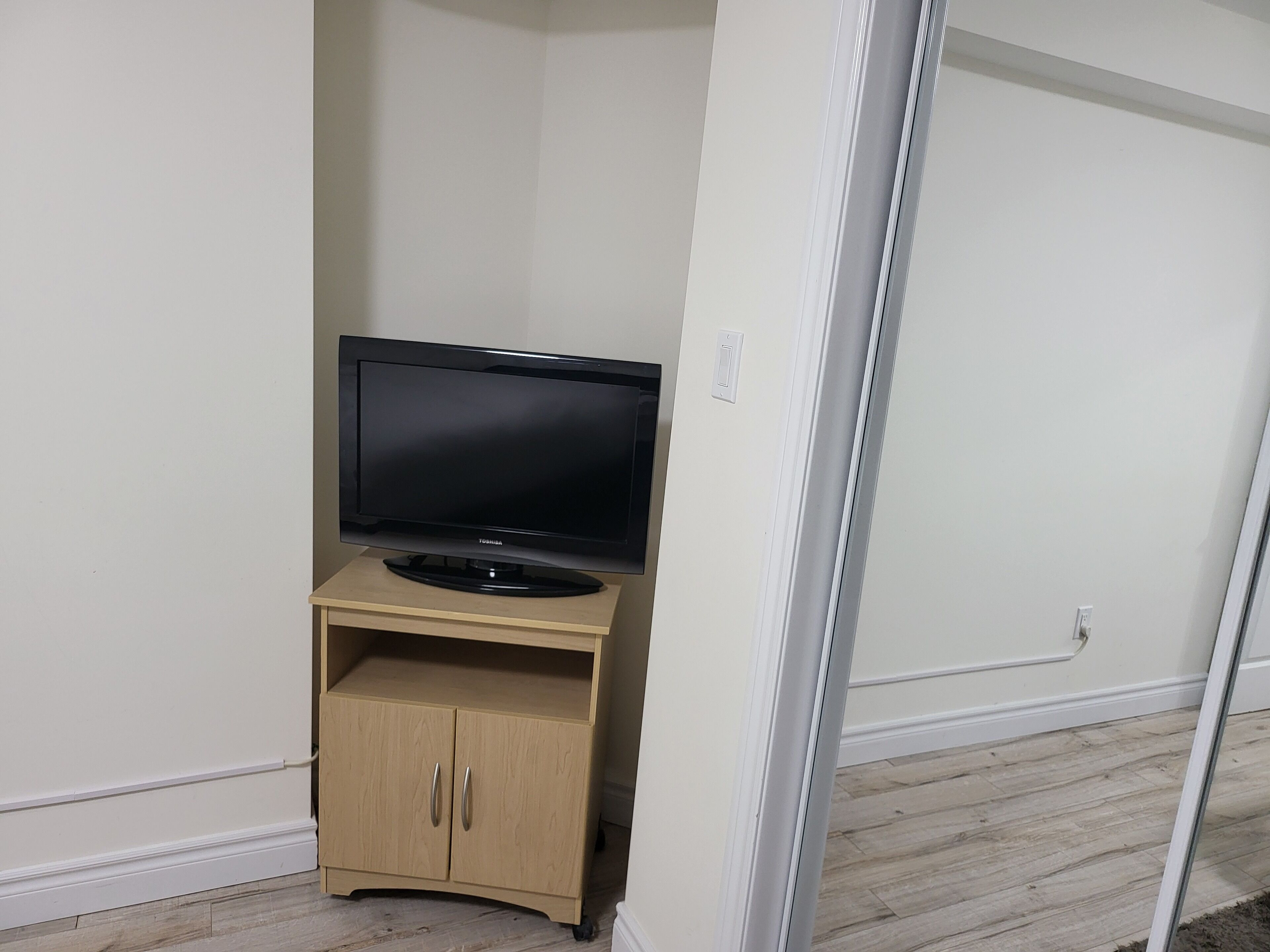A room with a flat-screen TV on a wooden stand, a white door, and a light-colored wooden floor.