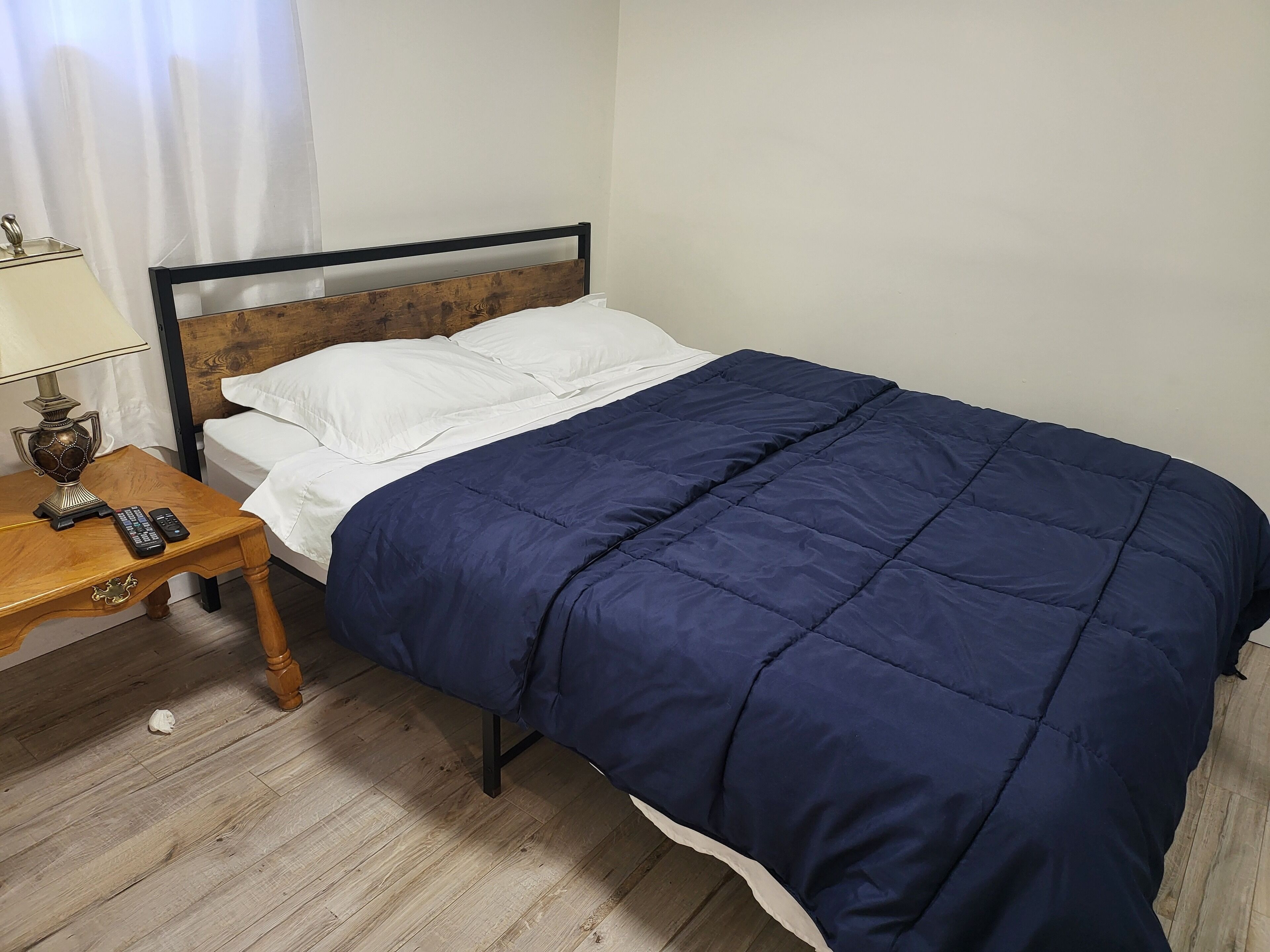 A neatly made bed with a dark blue bedspread and white pillows, a wooden bedside table with a lamp and remote control, and a wooden floor.