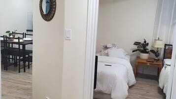 A hotel room with a bed, bedside table, lamp, and a small plant. There is a dining area with a table and chairs visible in the background.