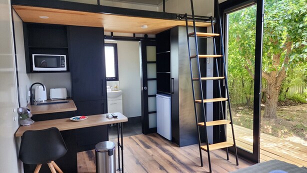 A compact kitchen with a microwave, toaster, and sink. A wooden ladder is leaning against the wall, and a small table with a chair is in the foreground. - Enchanting Tiny Home in Vibrant Dickson (Dickson)