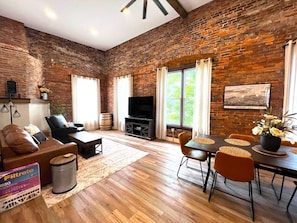 Una sala de estar con pared de ladrillo, chimenea, televisor, sofá, mesa de comedor con sillas y ventana con cortinas.