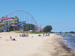 Una playa con una montaña rusa al fondo, gente relajándose en la arena y un cielo despejado.