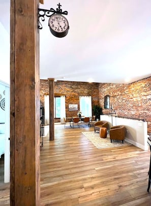 Amplia sala de estar con pared de ladrillo, piso de madera y un reloj colgado.