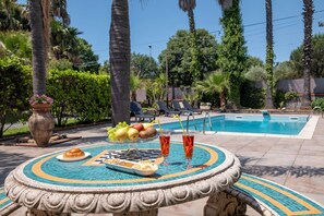 A poolside table with fruits, drinks, and a plate of food, surrounded by palm trees and a swimming pool.
