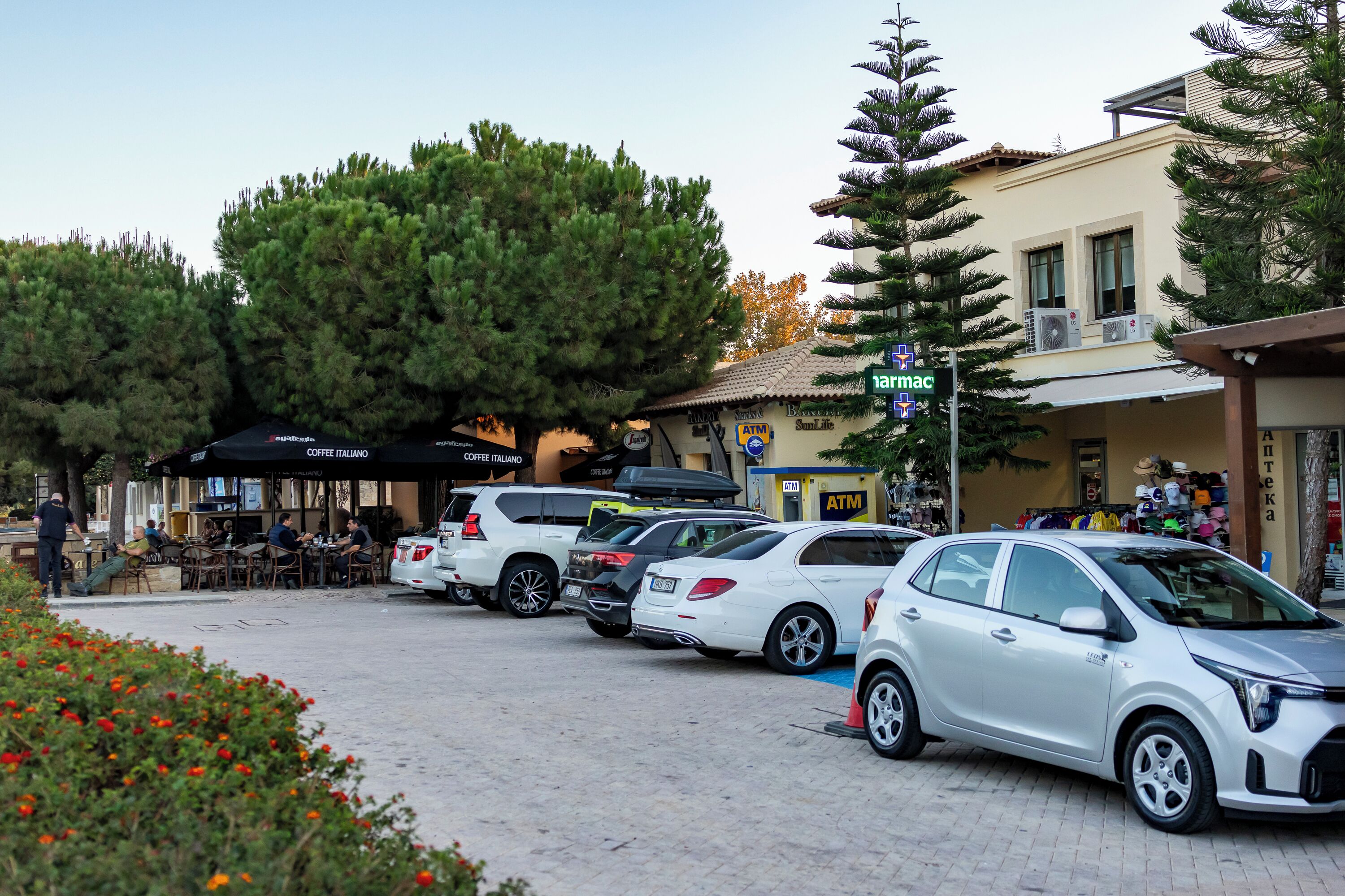A parking lot with several cars, outdoor seating at a cafe, and a building with a sign for 'ATM'.