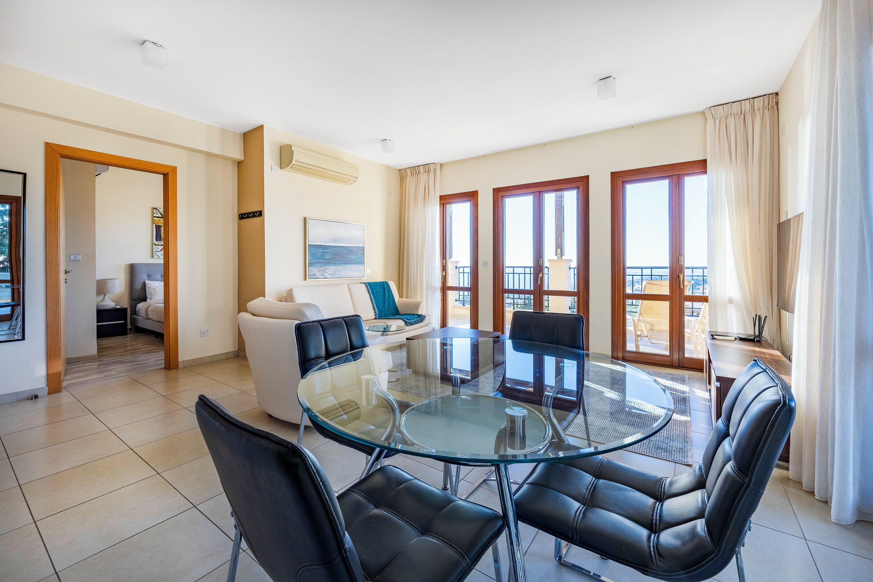 A spacious living area with a glass-top dining table surrounded by black leather chairs, a white sofa, and a balcony with a view.