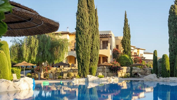 Una piscina con borde de piedra, rodeada de árboles bien recortados y un edificio con balcones.