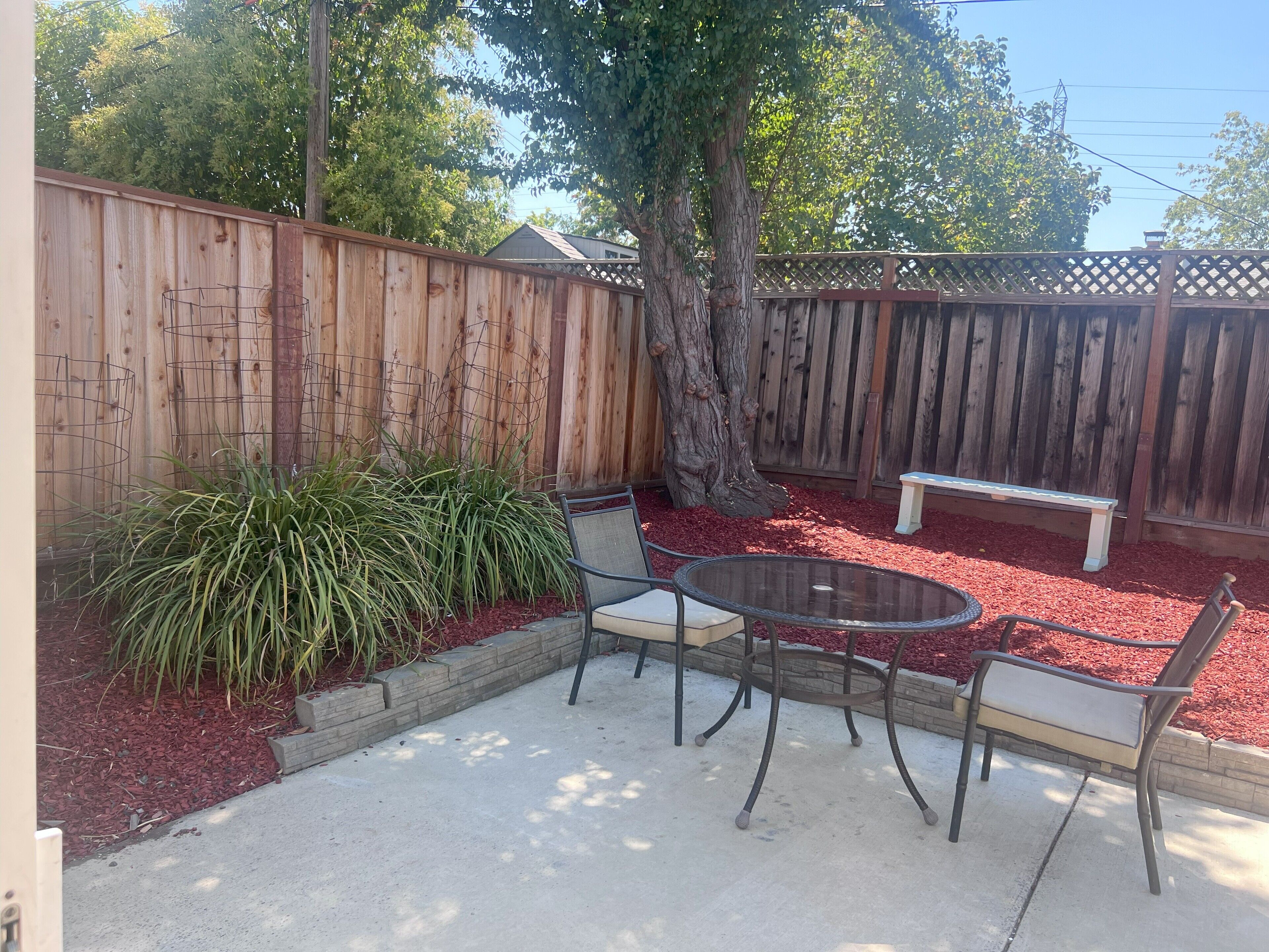 Een terras met een ronde glazen tafel en twee stoelen, omringd door rode mulch, met een houten schutting en een boom op de achtergrond.