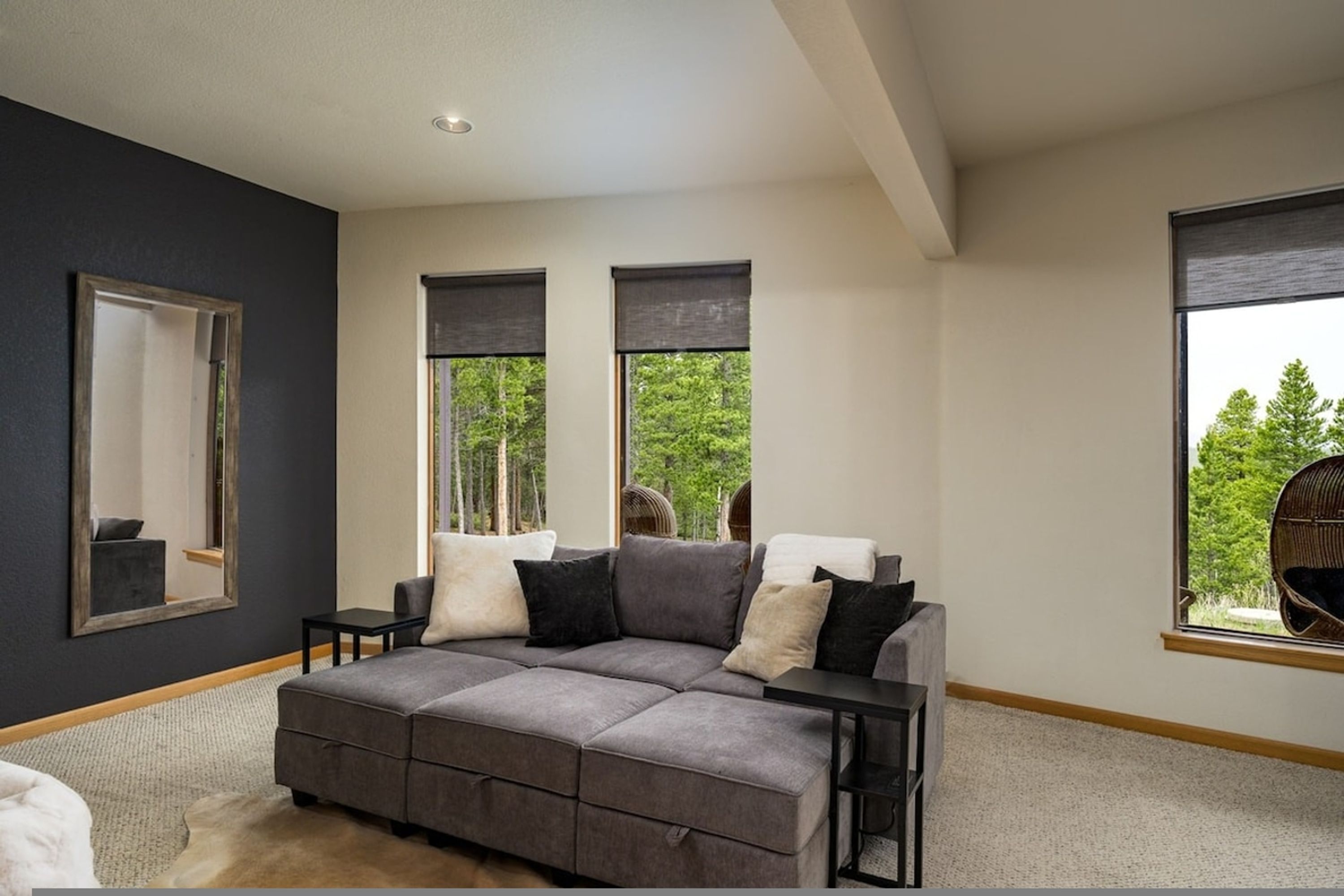 A modern living room with a sectional sofa, a large mirror, and two windows with blinds.