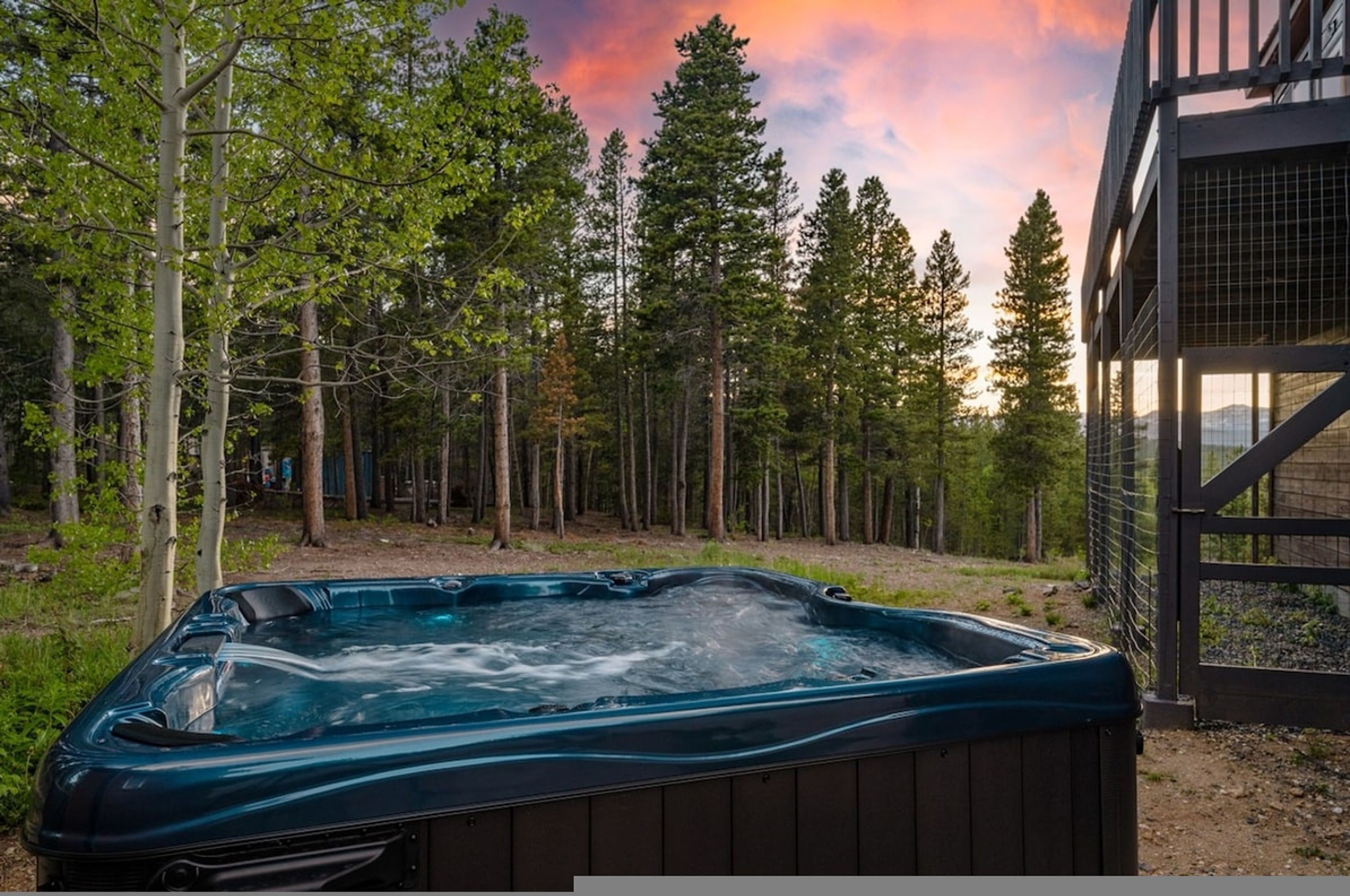 A hot tub in a forested area with a wooden deck and a metal fence.