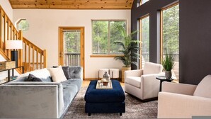A spacious living room with a wooden ceiling, large windows offering a view of the outdoors, a grey sofa, a blue ottoman, and a beige armchair. - Serene Retreat Amazing Views Hottub Sauna Xbox (Black Hawk)