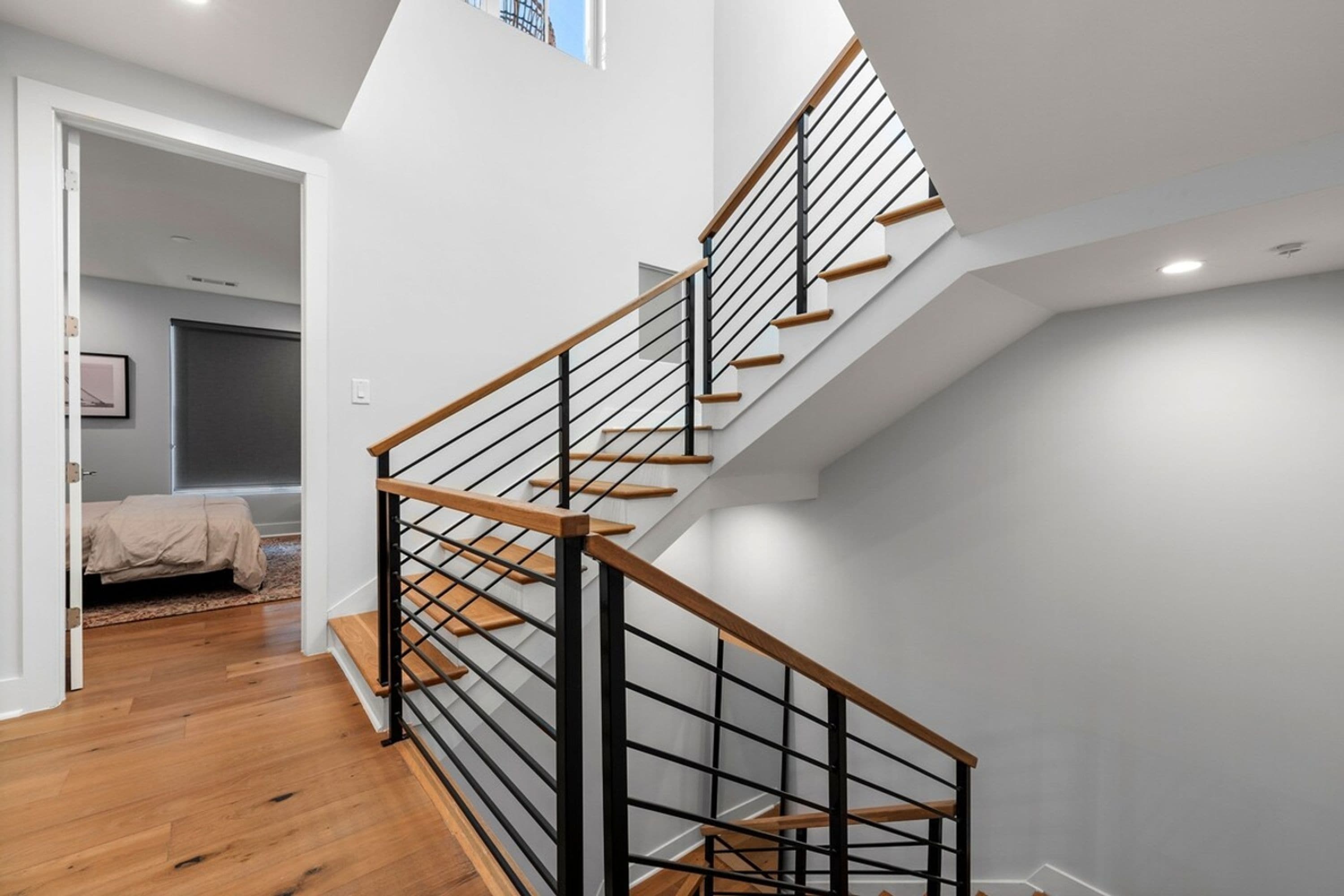 A modern staircase with wooden steps and black railings, leading to a bedroom with a bed and a window.