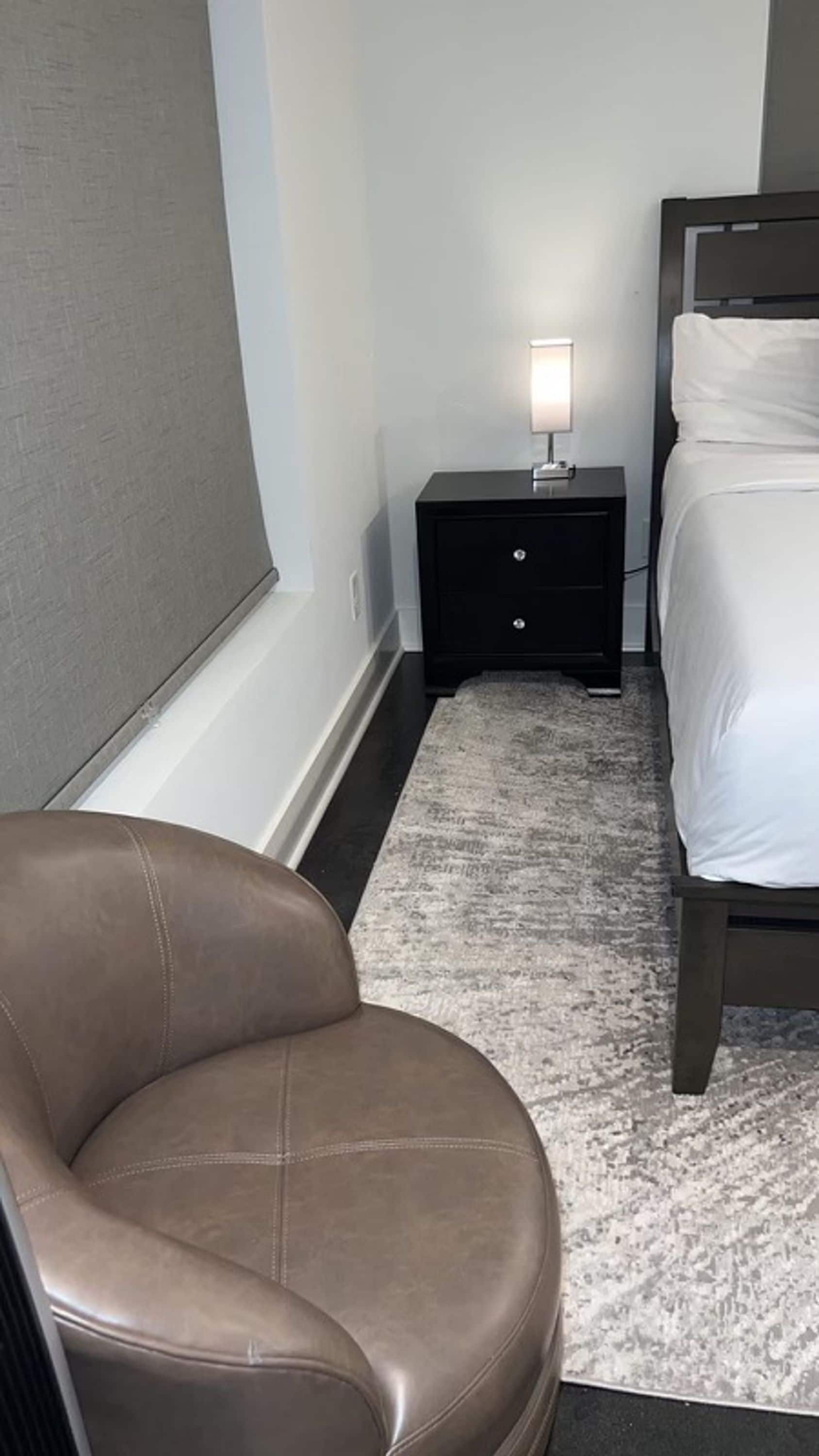 Une chambre à coucher avec un lit, une table de chevet avec une lampe, une chaise et un sol recouvert d’une moquette.