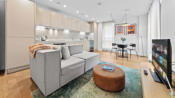 Ein modernes Wohnzimmer mit einem grauen Sofa, einem runden Hocker und einem Couchtisch. Es gibt einen Essbereich mit Tisch und Stühlen, und im Hintergrund eine Küche.
