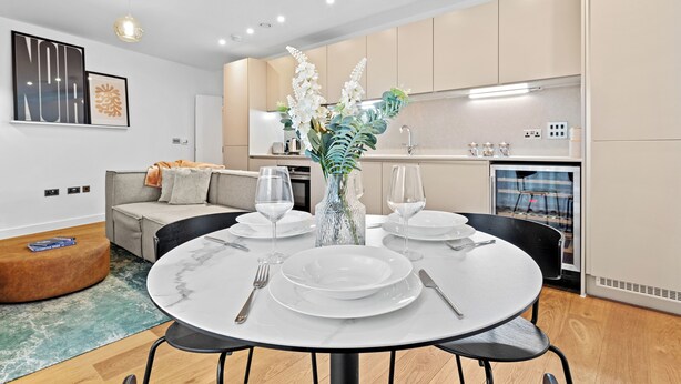 A modern dining area with a round table set for four, surrounded by a neutral-toned kitchen and a cozy seating area. - One Bed Ground Floor (London)