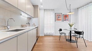 A modern kitchen with a large island, white cabinets, and a wooden floor. There is a dining area with a table and chairs, and a window with sheer curtains. - One Bed Ground Floor (London)