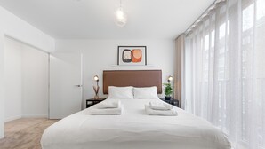 A modern hotel room with a large bed, a headboard, a bedside table with a lamp, and a window with sheer curtains. - Split Level Apartment (London)