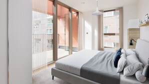 Une chambre moderne avec un grand lit, une porte-fenêtre coulissante donnant sur un balcon et une vue sur un immeuble en briques.