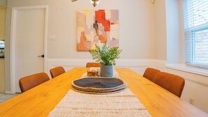 A dining area with a wooden table, chairs, a potted plant, and a wall-mounted abstract painting. - Spacious 4-bedroom 2-bath POOL HOUSE fully fenced (Spring)