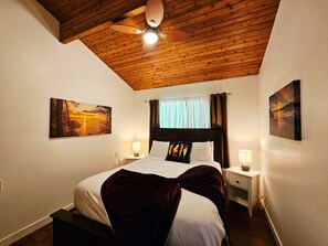 Ein Schlafzimmer mit Holzbalkendecke, einem Bett mit dunklem Kopfteil, zwei Nachttischen mit Lampen und zwei Bildern an den Wänden.