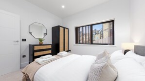 A modern bedroom with a large bed, a wardrobe, a mirror, and a window with a view of buildings.