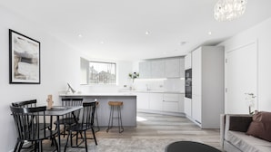 A modern kitchen with white cabinets, a dining area with black chairs, and a grey sofa. - Bejoux Court - Apt 1 (Harrow)