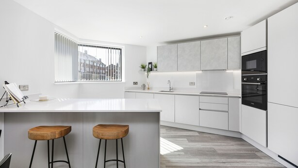 A modern kitchen with white cabinetry, a large island with stools, a built-in oven, and a window with blinds. - Bejoux Court - Apt 1 (Harrow)