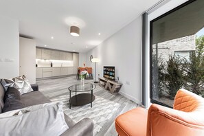 A modern living room with a sofa, armchair, and coffee table, a kitchen in the background, and a large window with a view of trees.