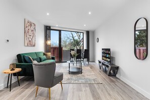 A modern living room with a green sofa, a grey armchair, a coffee table, and a TV stand with a bookshelf. - Bejoux Court - Apt 8 (Harrow)