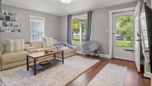 Ein Wohnzimmer mit einem beigen Sofa, einem Couchtisch, einem Sessel und einem Teppich. An den Wänden hängen Bilder und eine Tür führt nach draußen.