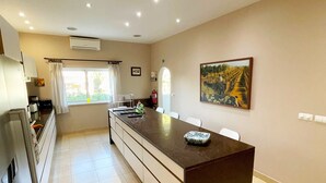 A modern kitchen with a central island, a dining area, and a large window providing natural light. - Villa Mandarin (Carvoeiro)