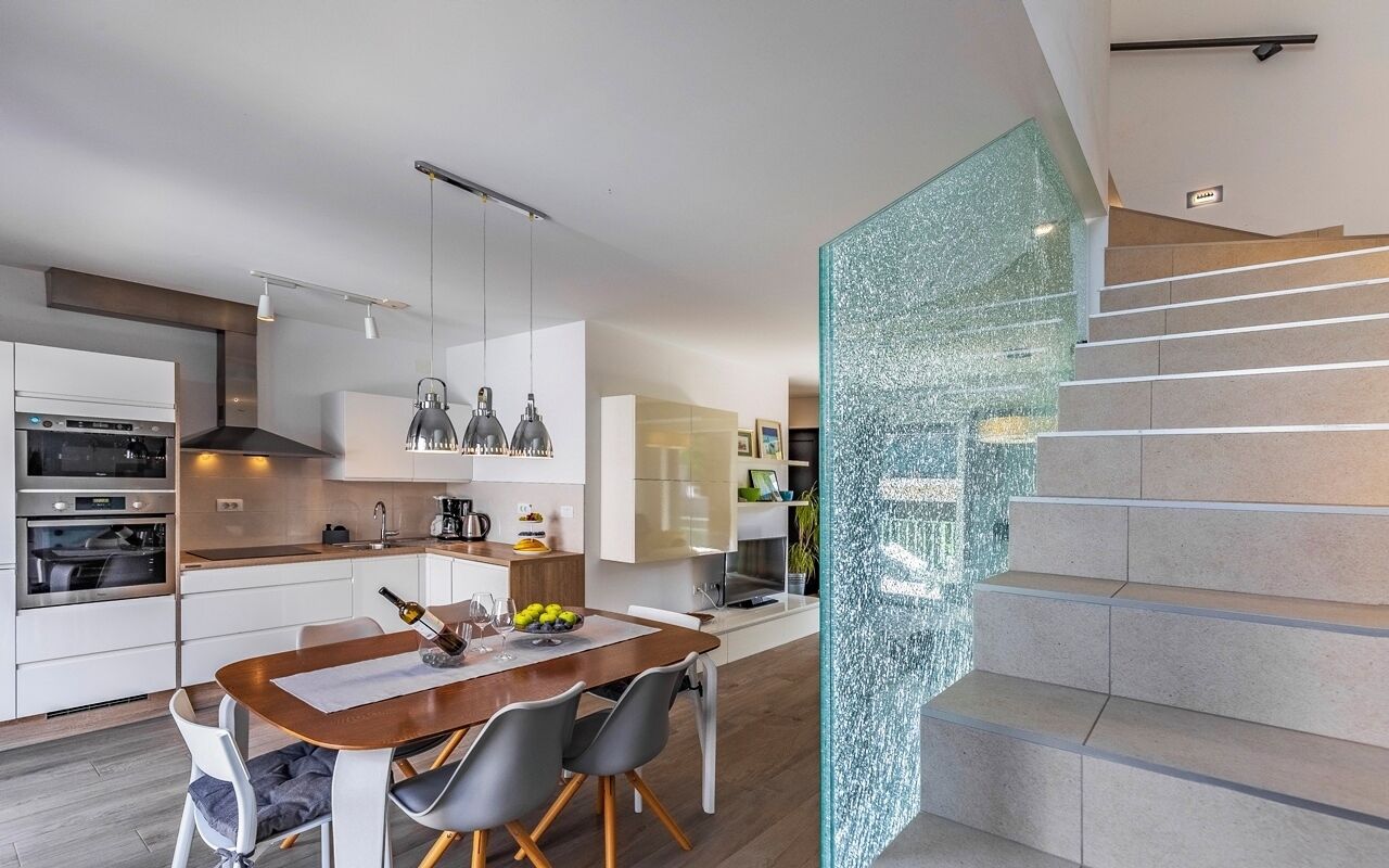 A modern kitchen with a dining area, a staircase with a glass railing, and a view of the living area.