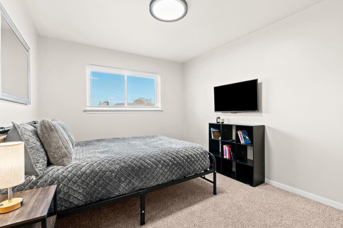 Une chambre à coucher avec un lit, une table de chevet avec une lampe, une bibliothèque et un téléviseur fixé au mur.
