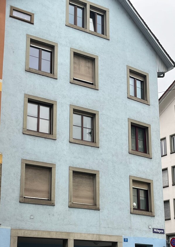 A multi-story building with a light blue facade, multiple windows with shutters, and a visible street sign indicating 'Mühlengasse'.